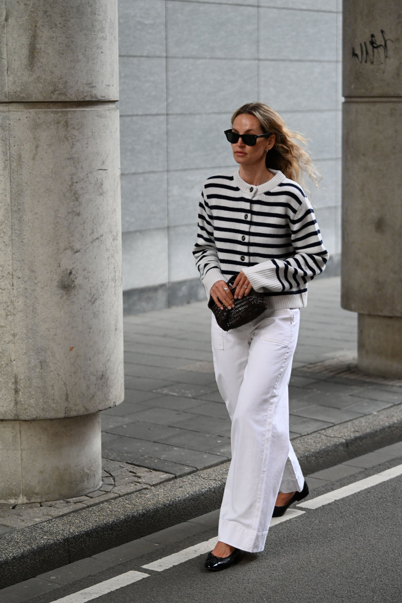 Eine Frau läuft eine Straße entlang. Sie trägt einen dunkelblau/weiß gestreiften Cardigan und eine weiße weite Jeans. In den Händen hält sie eine Tasche.