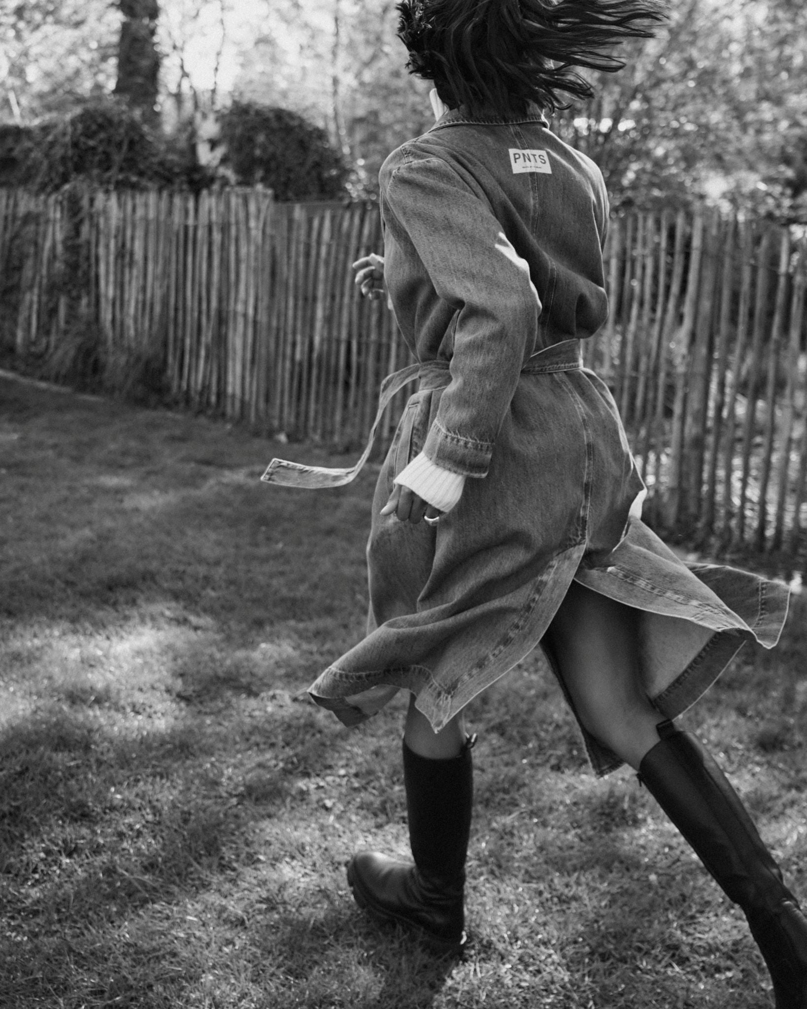 Eine Frau läuft über eine Wiese. Man sieht sie von hinten. Sie trägt kniehohe Stiefel und den Jeans Trenchcoat 27_THE SUIT UP TRENCH