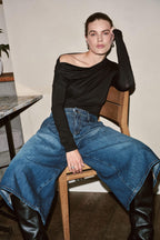 Eine Frau sitzt auf einem Holzstuhl in einem schwarzen asymmetrischen Top, einer weiten Jeans-Culotte und schwarzen Lederstiefeln.