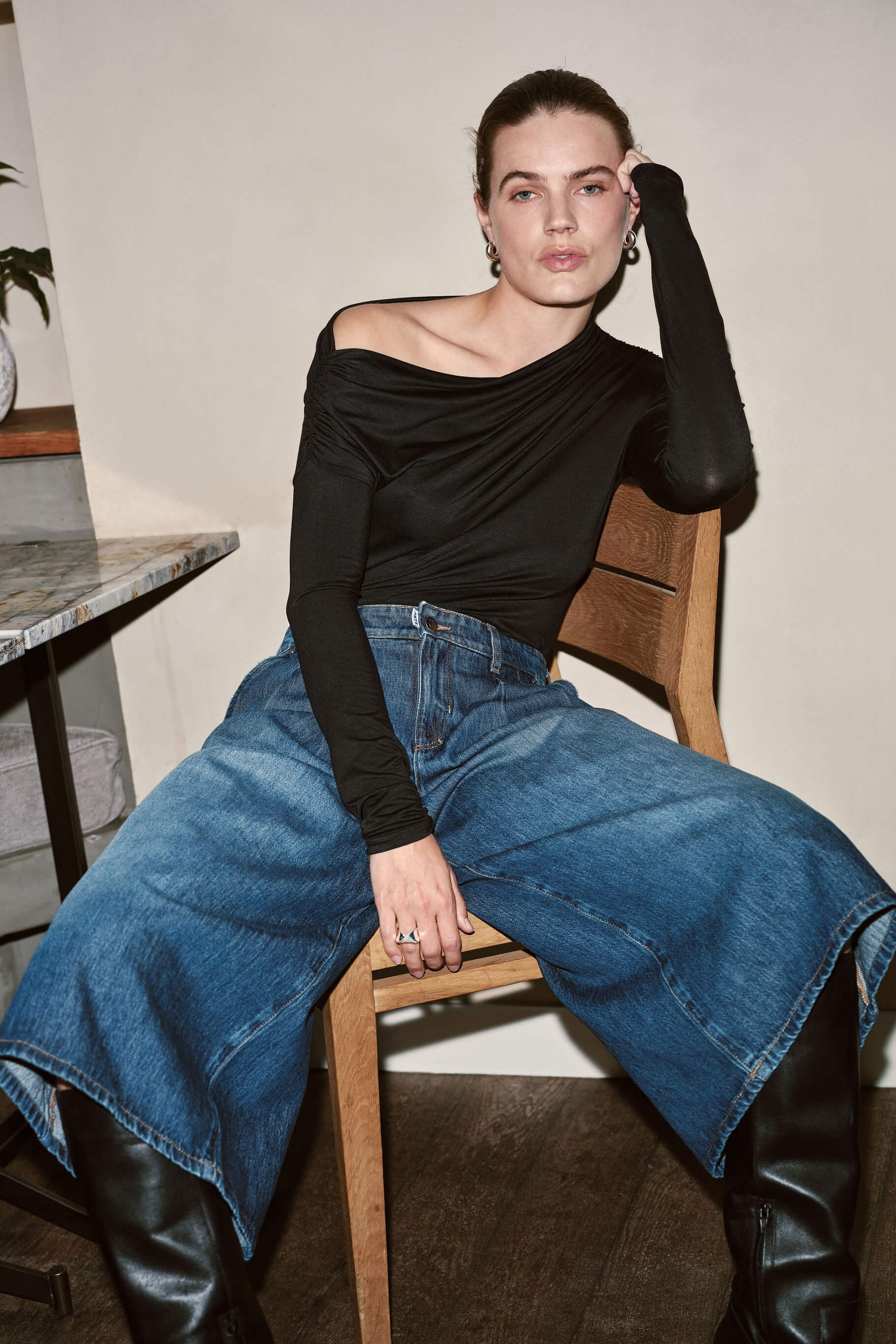 Eine Frau sitzt auf einem Holzstuhl in einem schwarzen asymmetrischen Top, einer weiten Jeans-Culotte und schwarzen Lederstiefeln.