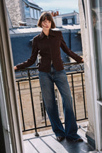 Frau in dunkelbrauner Zip-Jacke und weiter blauer Jeans auf einem Balkon vor Stadtkulisse.