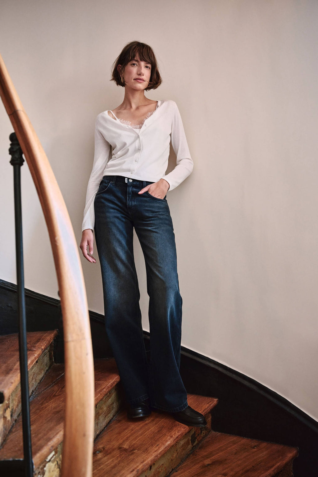 Frau in weißem Cardigan, Top mit Spitzenbesatz und dunkelblauer Wide-Leg-Jeans auf einer Holztreppe.
