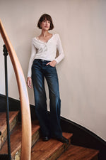 Frau in weißem Cardigan, Top mit Spitzenbesatz und dunkelblauer Wide-Leg-Jeans auf einer Holztreppe.