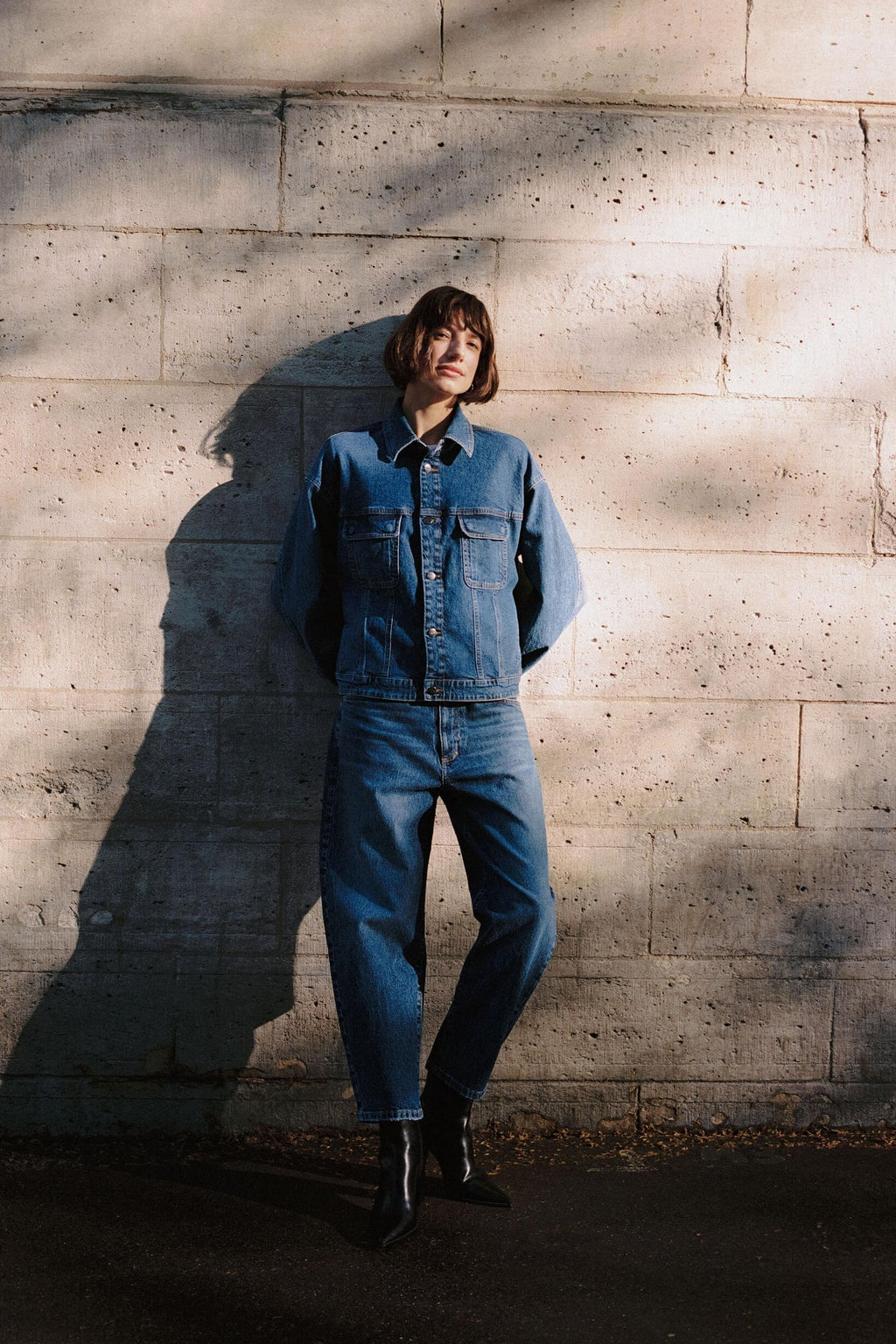 Frau in einem Denim-Outfit mit blauer Jeansjacke und passender Jeans vor einer hellen Steinmauer.