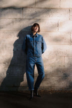 Frau in einem Denim-Outfit mit blauer Jeansjacke und passender Jeans vor einer hellen Steinmauer.