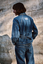 Rückansicht einer Frau in blauer Denim-Jacke mit Logo-Patch und Jeans vor einer hellen Steinwand.