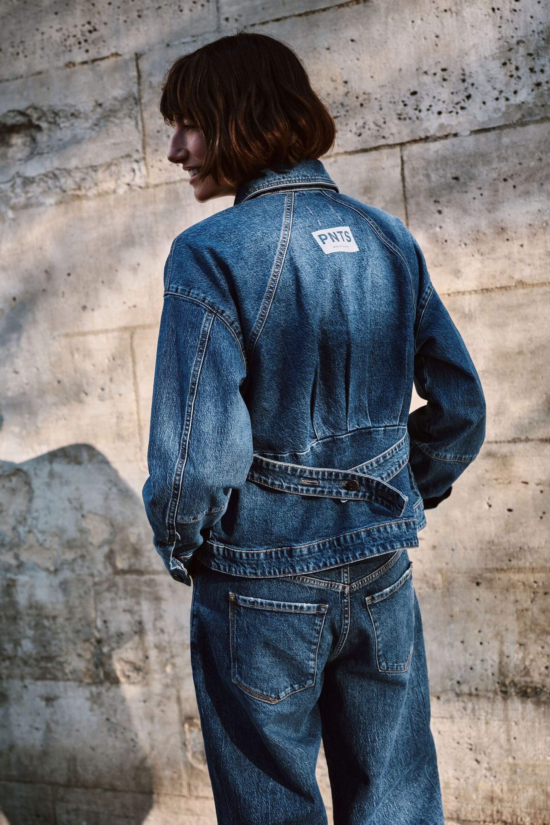 Rückansicht einer Frau in blauer Denim-Jacke mit Logo-Patch und Jeans vor einer hellen Steinwand.