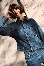 Lächelnde Frau mit Bob-Frisur in blauer Denim-Jacke und Jeans vor einer hellen Steinwand.