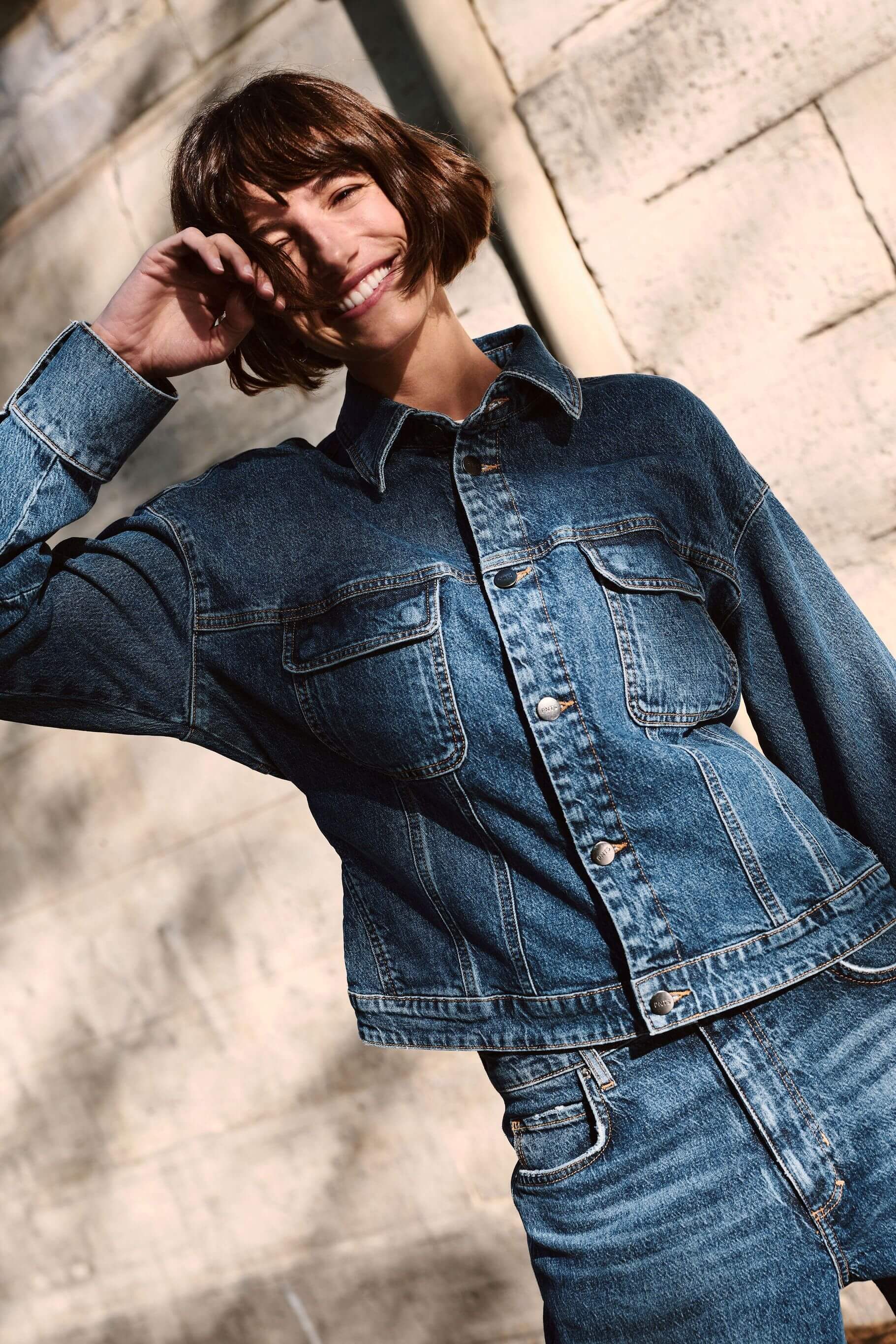 Lächelnde Frau mit Bob-Frisur in blauer Denim-Jacke und Jeans vor einer hellen Steinwand.