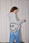 Rückansicht: Hellgrauer Oversize-Strick-Hoodie mit großem eingestricktem Smiley-Rückenmotiv, kombiniert mit lockerer Blue Jeans.