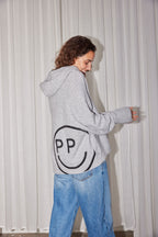 Rückansicht: Hellgrauer Oversize-Strick-Hoodie mit großem eingestricktem Smiley-Rückenmotiv, kombiniert mit lockerer Blue Jeans.