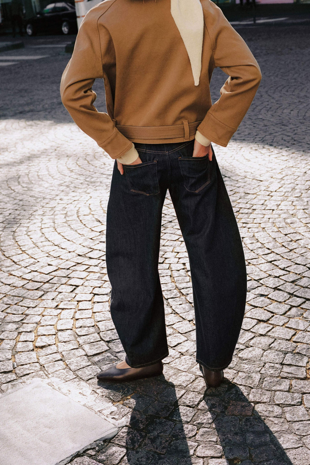 Rückansicht einer Frau in kamelfarbener Jacke und dunkelblauer Wide-Leg-Jeans auf Kopfsteinpflaster.