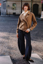 Frau in kamelfarbener Kurzjacke mit Gürtel und dunkelblauer Wide-Leg-Jeans auf einer Kopfsteinpflasterstraße.