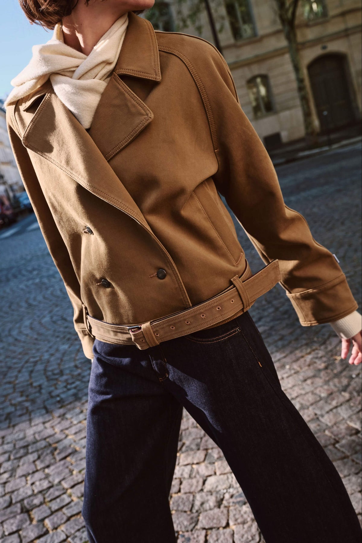 Frau in kamelfarbener Kurzjacke mit Gürtel und dunkelblauer Jeans auf Kopfsteinpflaster.