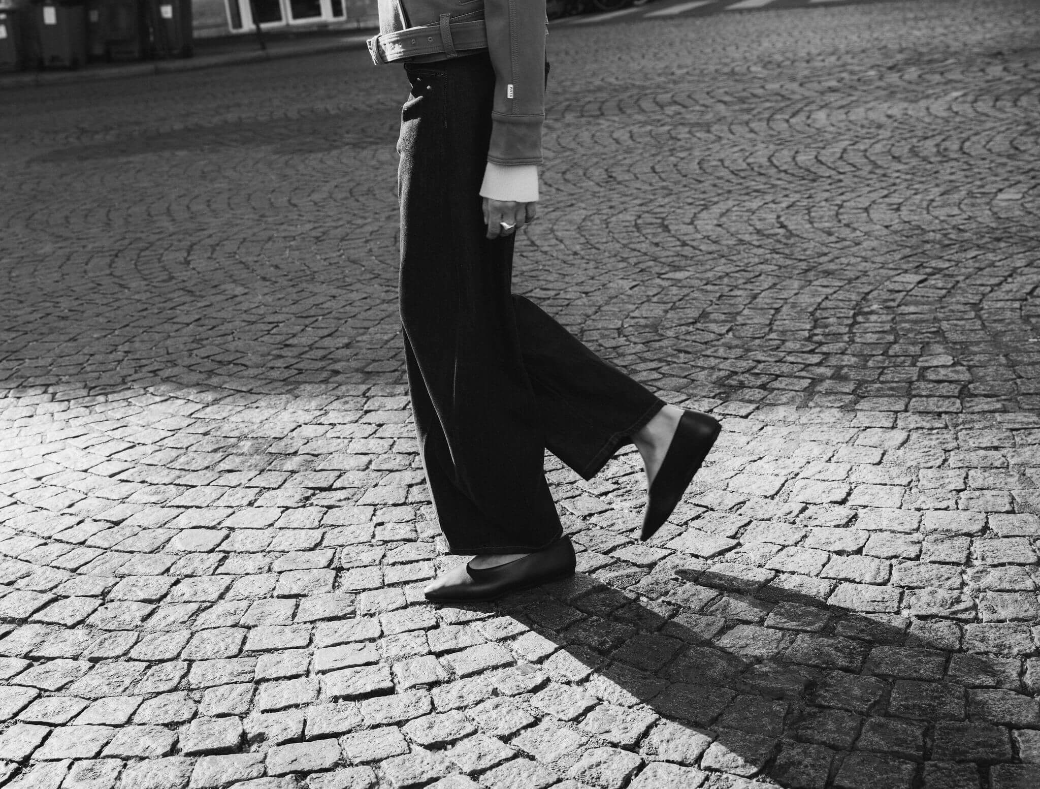 Schwarz-Weiß-Aufnahme einer Person in weiter Hose und flachen Schuhen beim Gehen auf Kopfsteinpflaster.