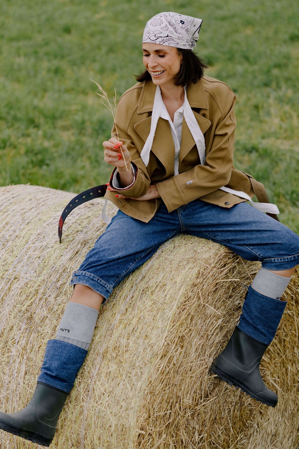 Outdoor-Trageansicht: Kurzer camel-brauner Trenchcoat/Jacke mit Bindegürtel, getragen auf einem Strohballen im modernen Country-Look.