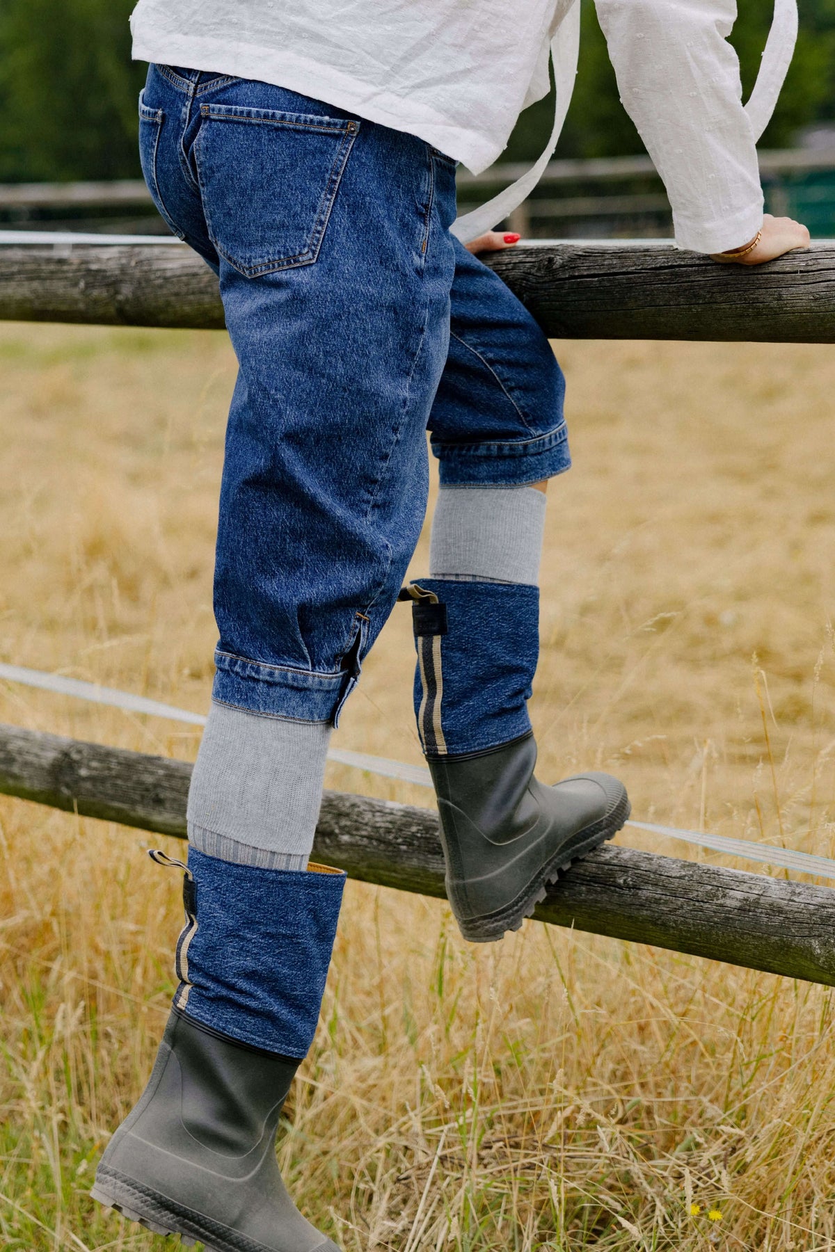 Outdoor-Trageansicht: Mittelblaue Damen-Jeans im verkürzten Schnitt mit Saumschlitz, kombiniert mit grauen Kniestrümpfen und Outdoor-Stiefeln an einem Holzzaun.