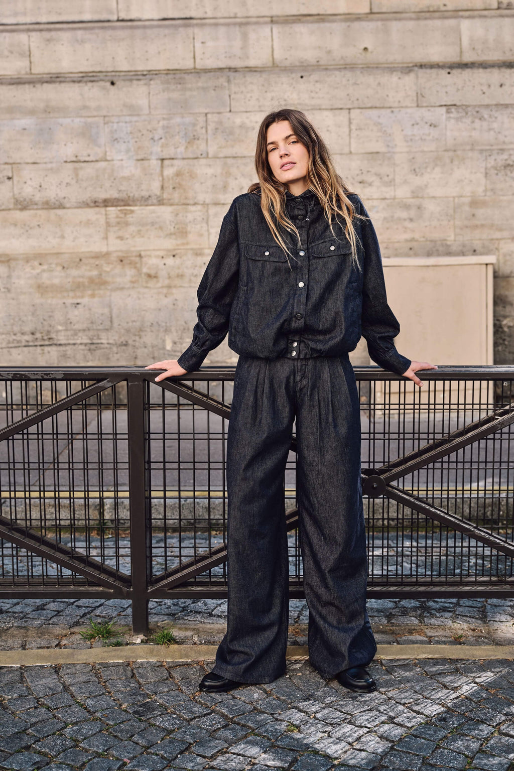 Frau in einem dunklen Denim-Outfit mit Jacke und weit geschnittener Hose, stehend an einem Metallzaun vor einer Steinwand.