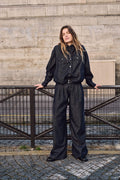 Frau in einem dunklen Denim-Outfit mit Jacke und weit geschnittener Hose, stehend an einem Metallzaun vor einer Steinwand.