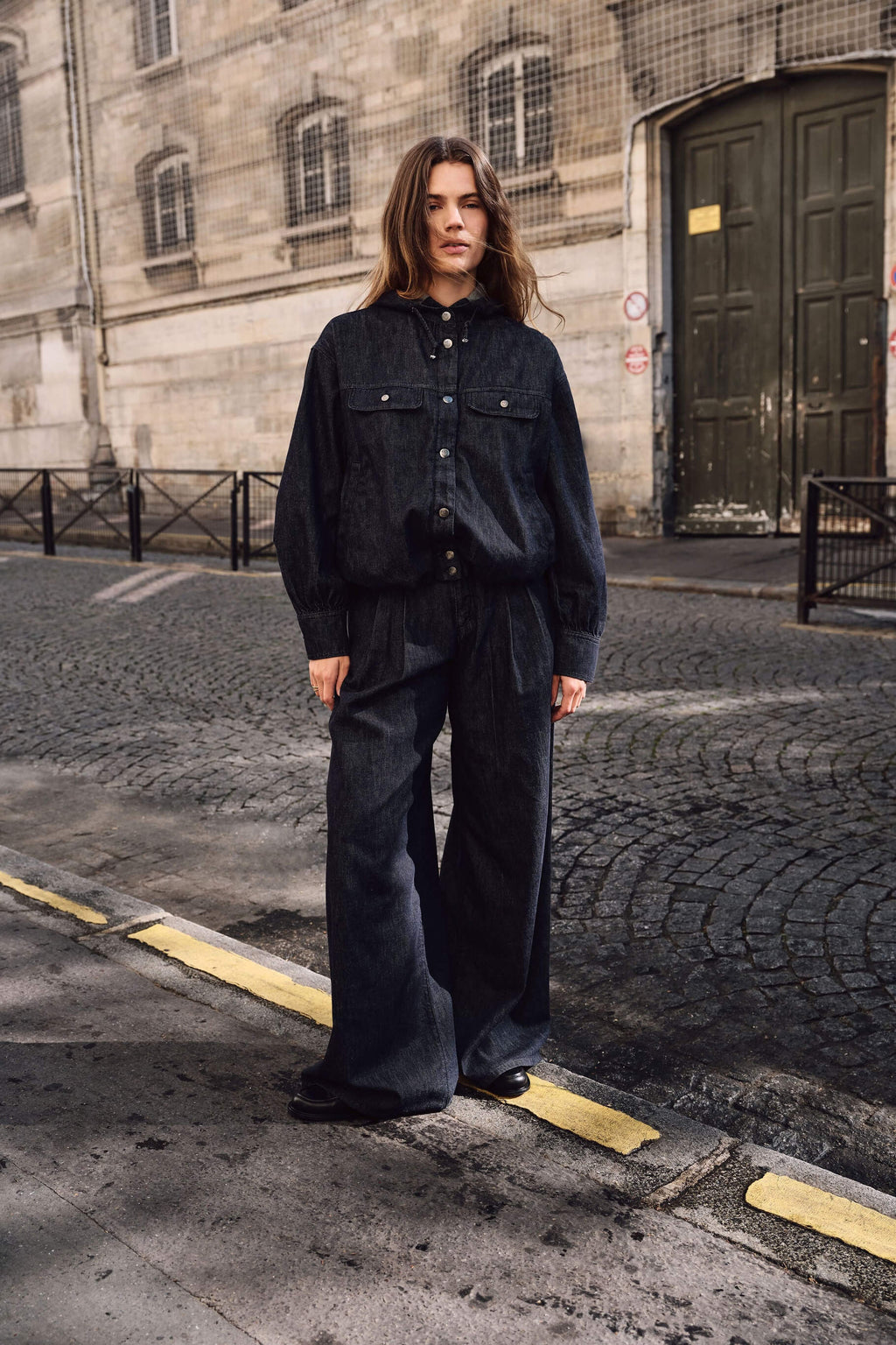 Frau in einem dunklen Denim-Outfit mit Kapuzenjacke und weiter Hose auf einer Kopfsteinpflasterstraße.
