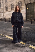 Frau in einem dunklen Denim-Outfit mit Kapuzenjacke und weiter Hose auf einer Kopfsteinpflasterstraße.