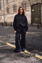 Frau in einem dunklen Denim-Outfit mit Kapuzenjacke und weiter Hose auf einer Kopfsteinpflasterstraße.