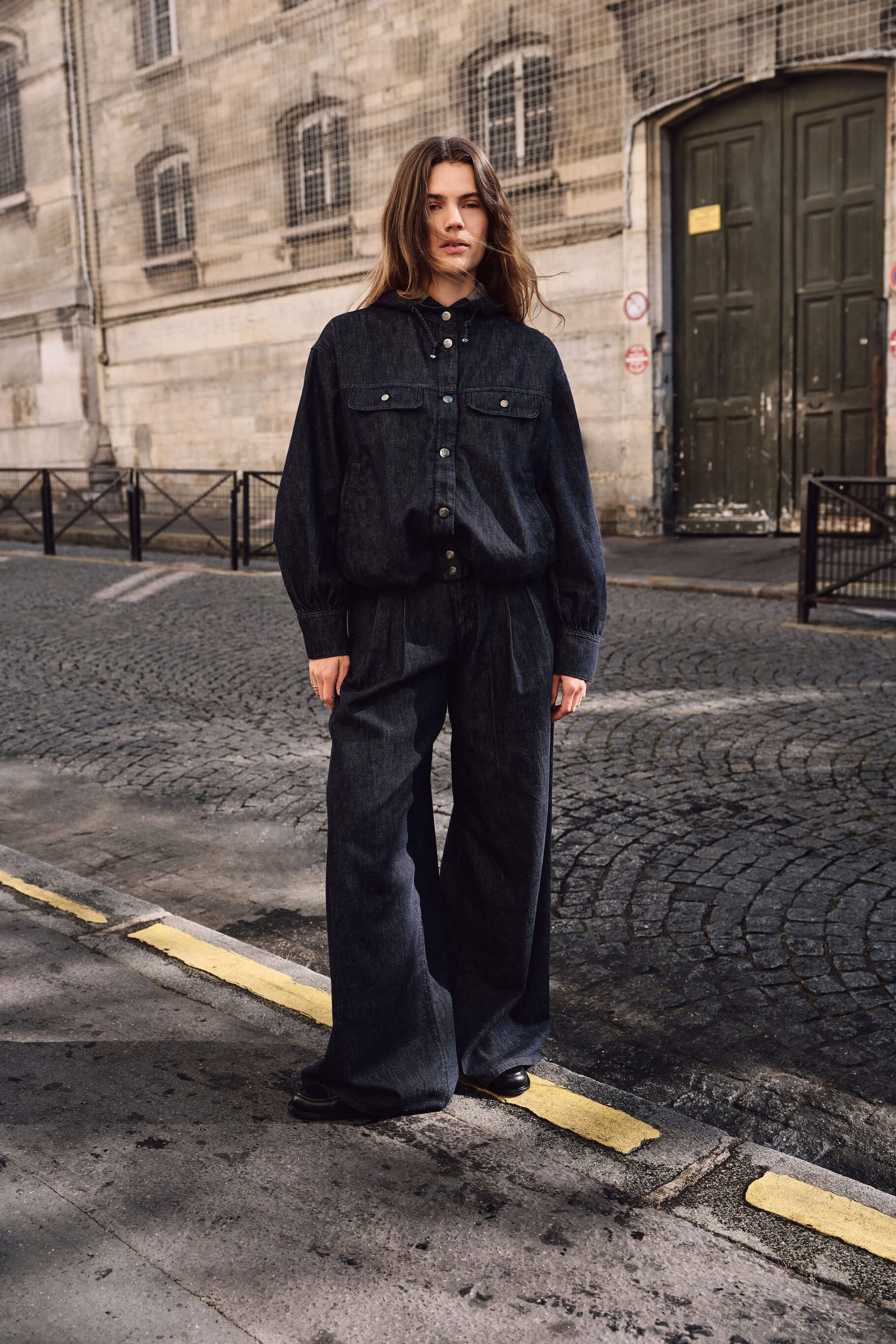 Frau in einem dunklen Denim-Outfit mit Kapuzenjacke und weiter Hose auf einer Kopfsteinpflasterstraße.