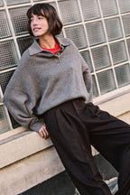 Frau in grauem Polo-Strickpullover, rotem Akzent und dunkelbrauner Wide-Leg-Hose vor einer Wand aus Glasbausteinen.
