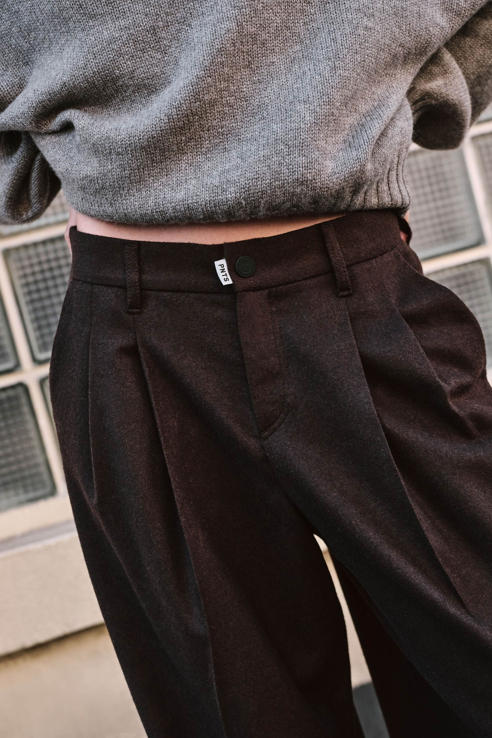 Detailansicht einer dunkelbraunen Bundfaltenhose mit weißem Logo-Label am Bund und grauem Strickpullover.