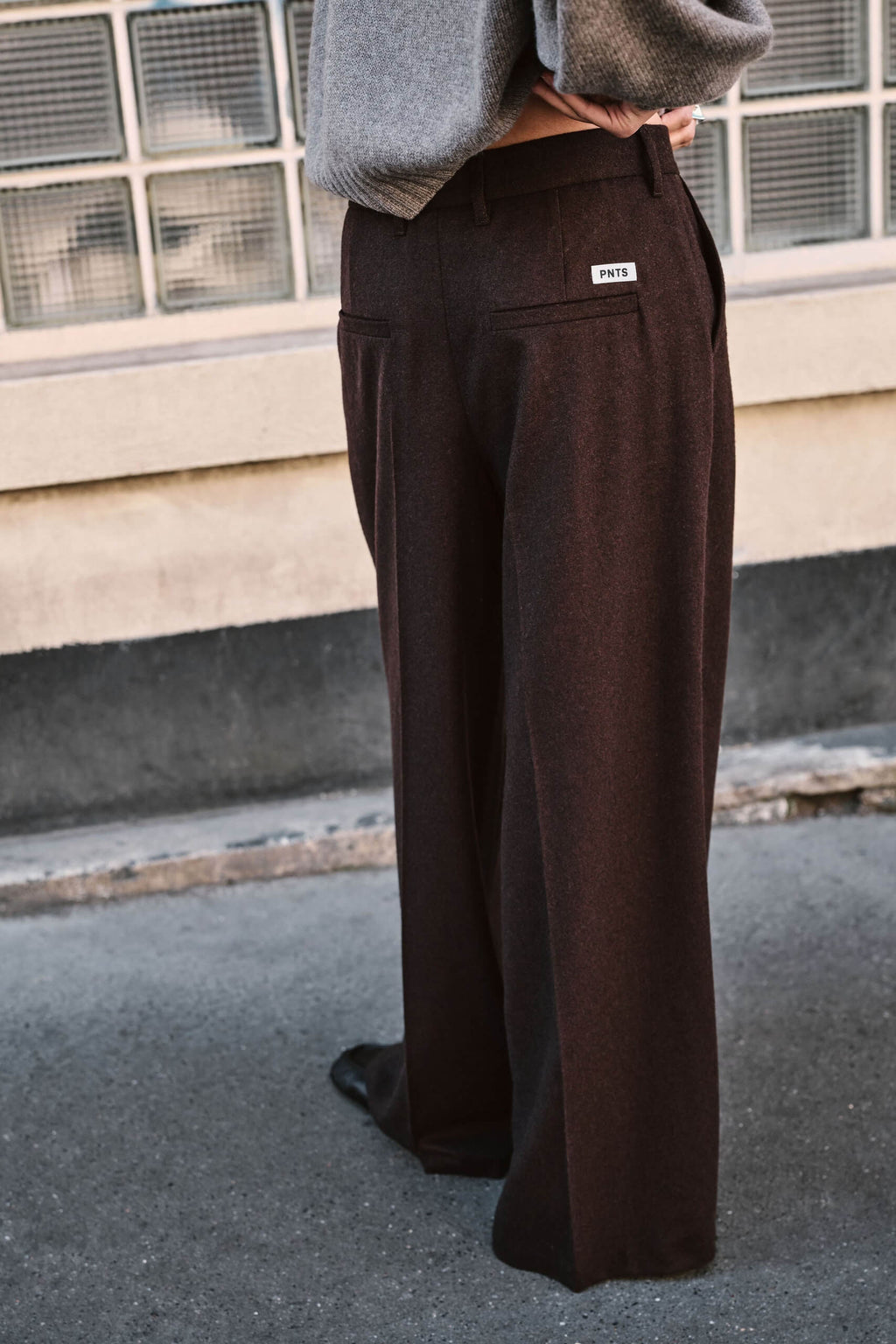 Rückansicht einer dunkelbraunen Wide-Leg-Hose mit weißem Logo-Label an der Gesäßtasche und grauem Pullover.