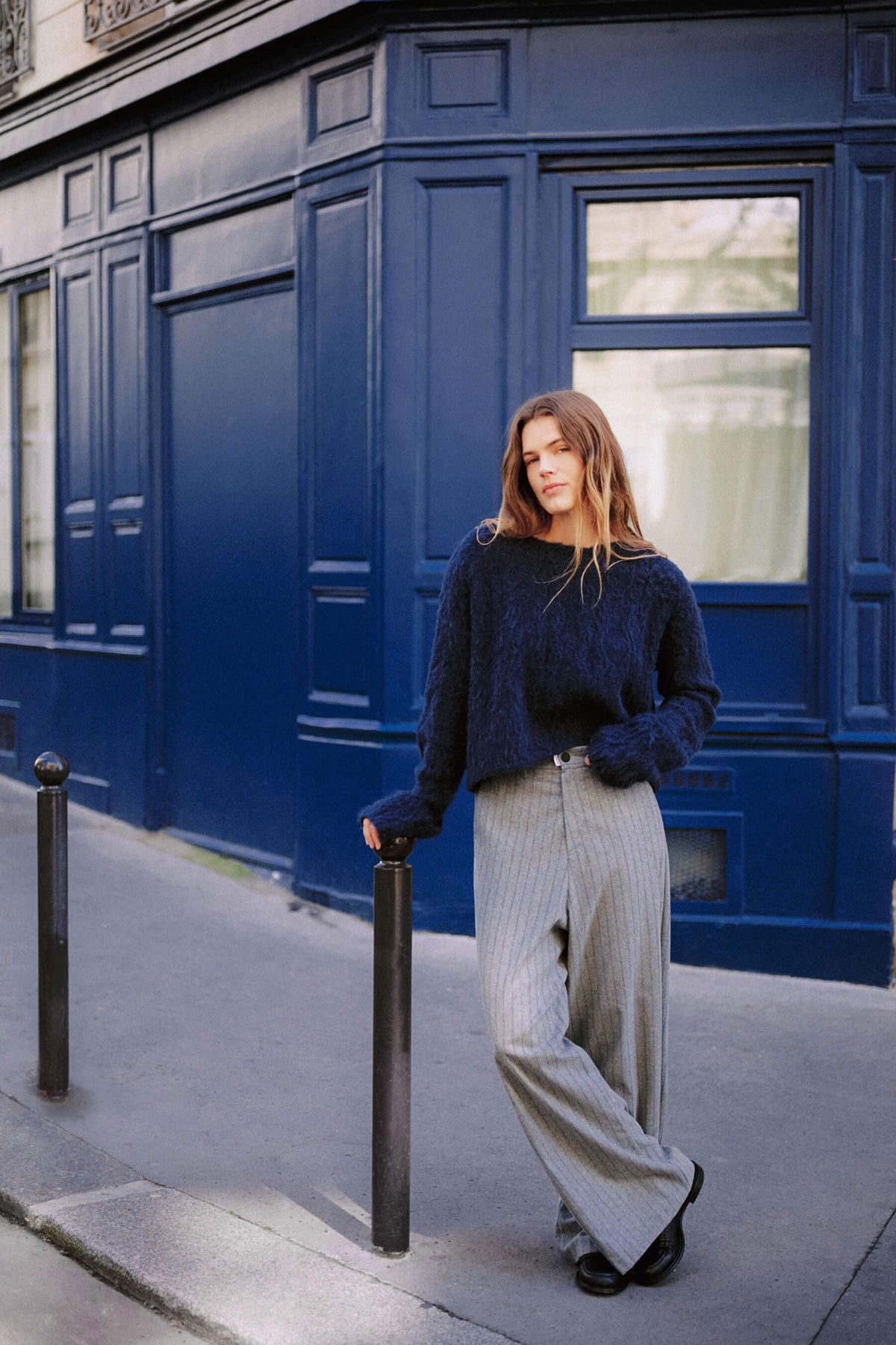 Frau in dunkelblauem Flausch-Pullover und grauer Wide-Leg-Hose mit feinem Muster vor einer blauen Hausfassade.
