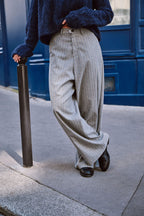 Detailansicht einer grauen Wide-Leg-Hose mit feinem Streifenmuster und schwarzen Loafern vor blauem Hintergrund.