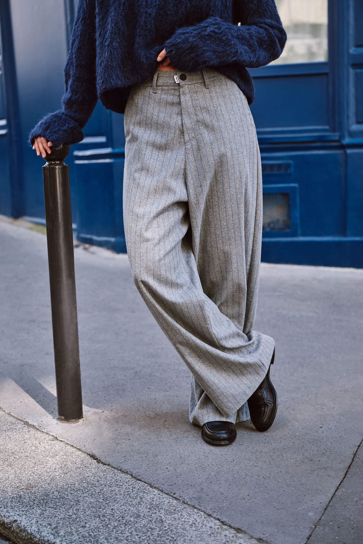 Detailansicht einer grauen Wide-Leg-Hose mit feinem Streifenmuster und schwarzen Loafern vor blauem Hintergrund.