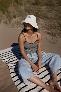 Frau sitzt auf einem gestreiften Handtuch am Strand mit einer Mütze auf. Sie trägt die Hose "05_THE O SHAPE EXTREME"