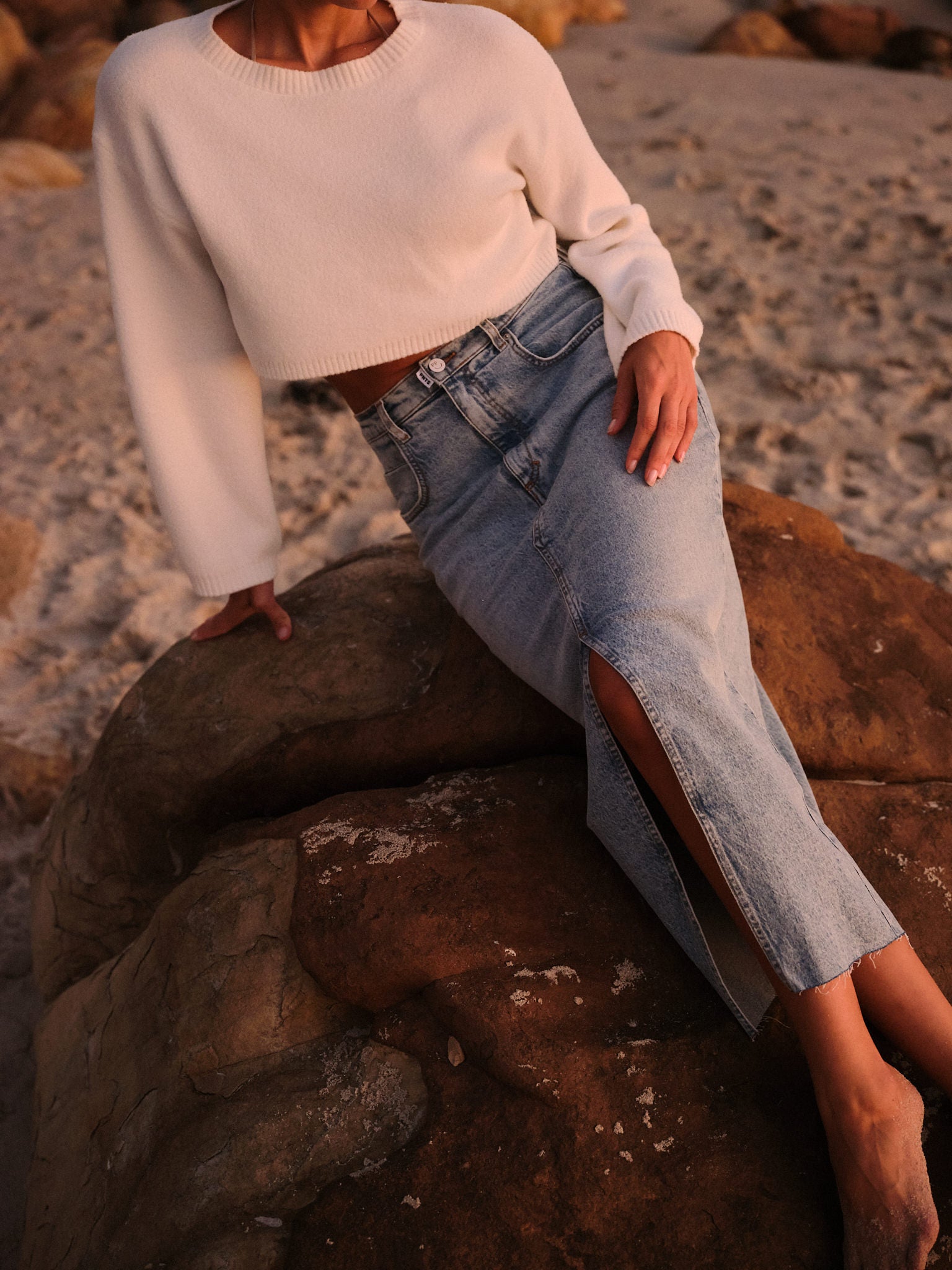 Frau sitzt mit einem weißen cropped Pullover auf einem Stein am Strand. Sie trägt den Rock "15_LETS ROCK" 
