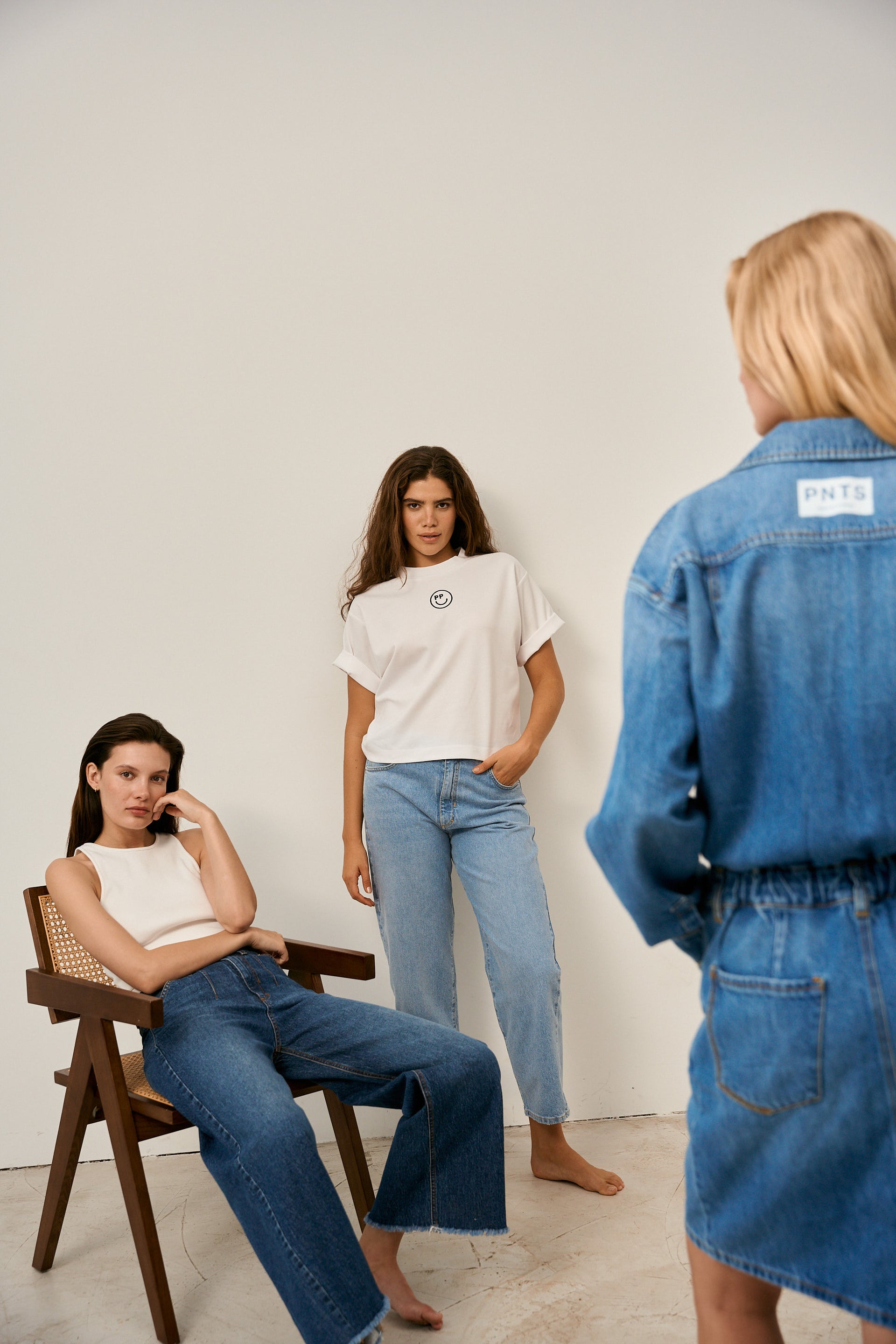 Es sind drei Frauen in dem Bild zu sehen. Eine sitzt auf einem Stuhl, eine steht an einer Wand und die andere steht mit dem Rücken zur Kamera.