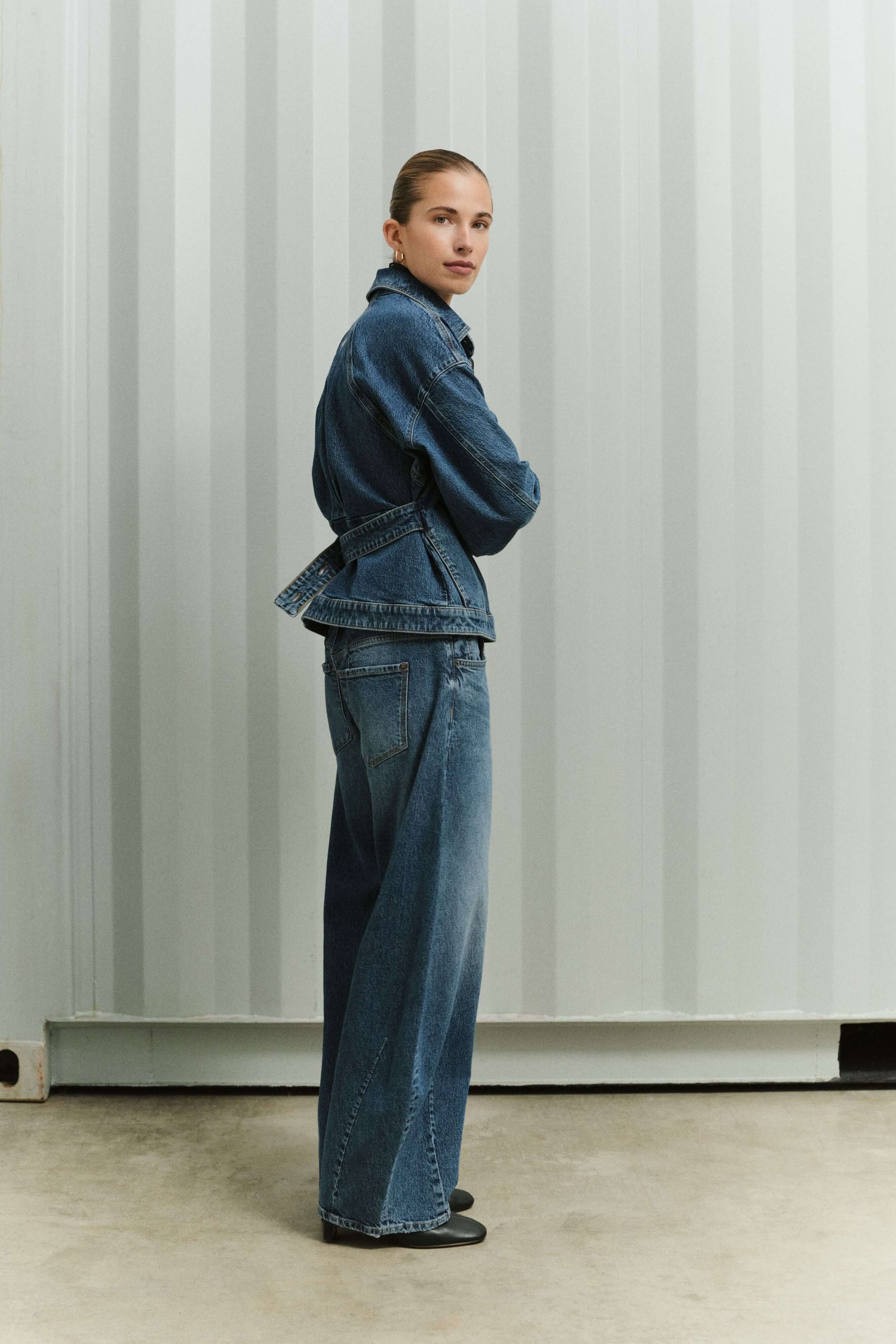 Frau in einer taillierten Jeansjacke mit Gürtel und weit geschnittener Jeans in der Seitenansicht.