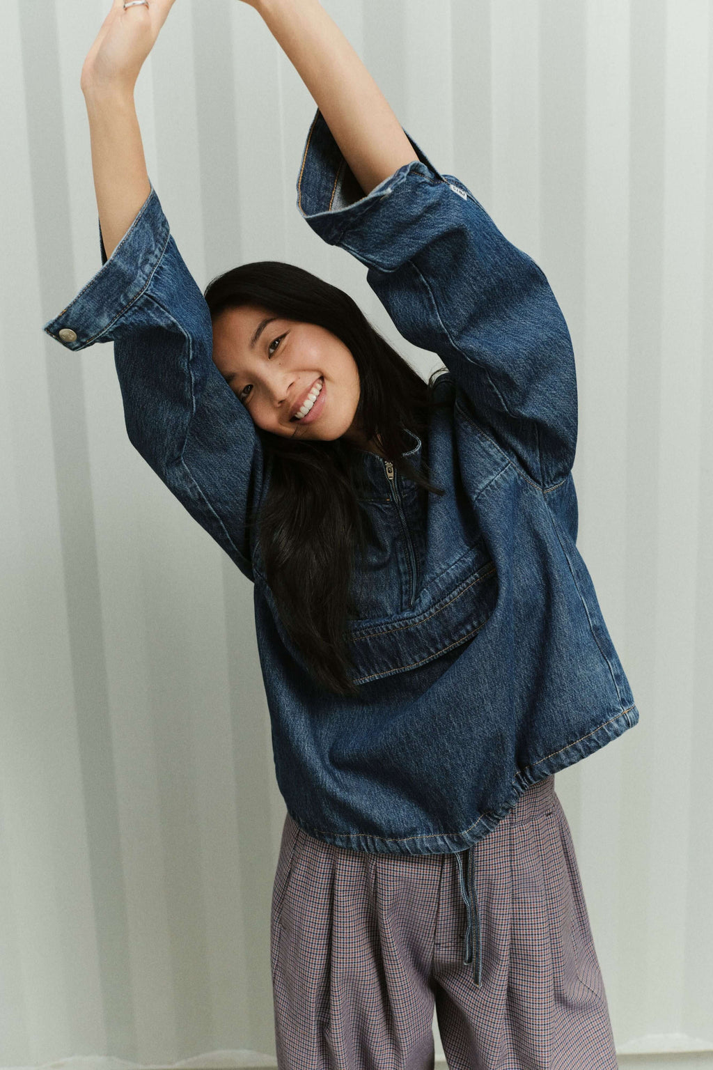 Eine lächelnde Frau mit erhobenen Armen in einem blauen Denim Sip Blouse und einer fein gemusterten Hose vor gestreiftem Hintergrund.