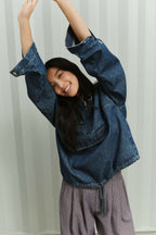 Eine lächelnde Frau mit erhobenen Armen in einem blauen Denim Sip Blouse und einer fein gemusterten Hose vor gestreiftem Hintergrund.