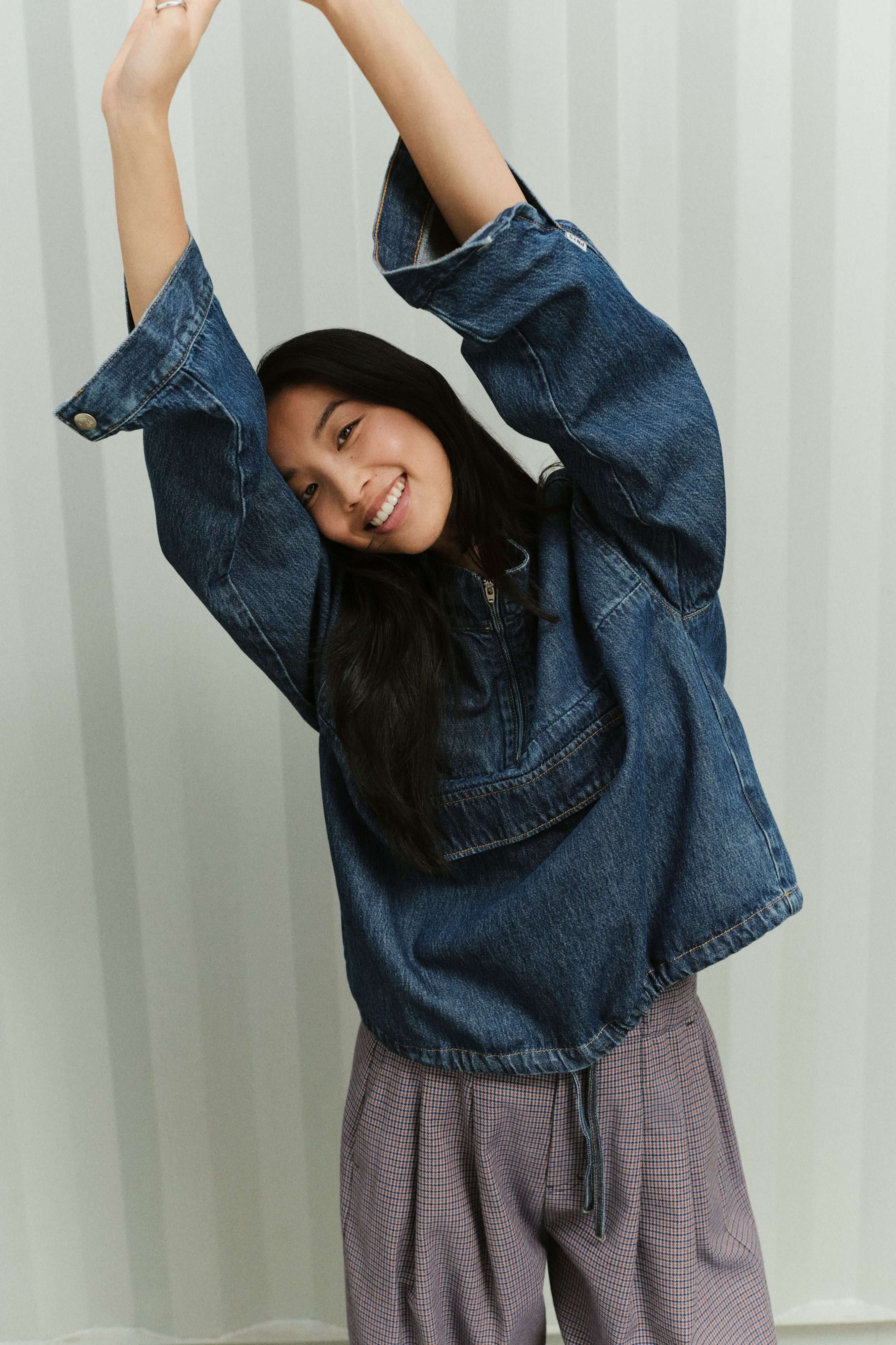 Eine lächelnde Frau mit erhobenen Armen in einem blauen Denim Sip Blouse und einer fein gemusterten Hose vor gestreiftem Hintergrund.