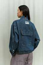 Rückansicht eines blauen Denim-Oberteil mit weißem PNTS-Label und einer großen Tasche auf dem Rücken.