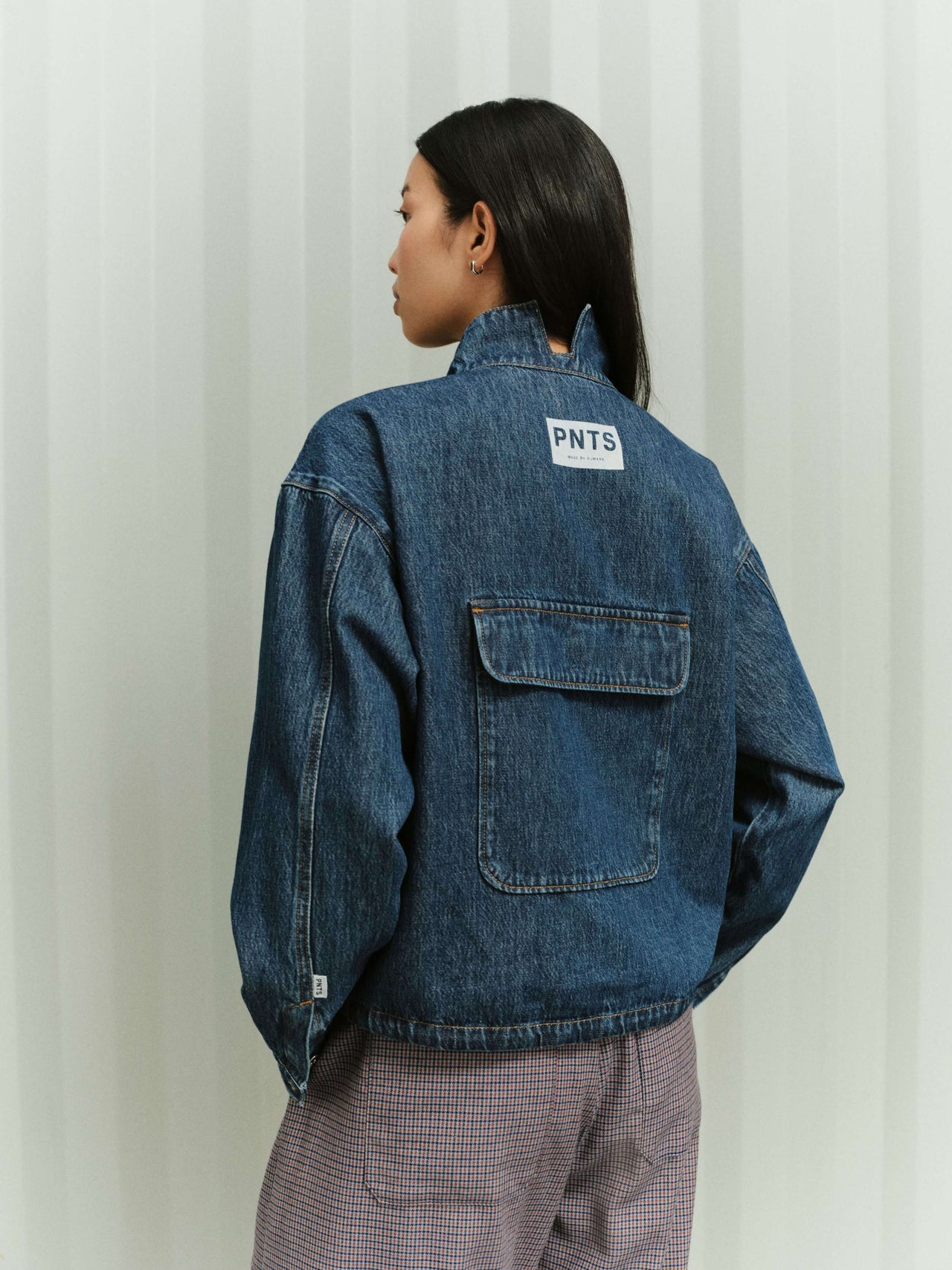 Rückansicht einer Frau in einer blauen Denim-Jacke mit Logo-Patch und einer karierten Hose.