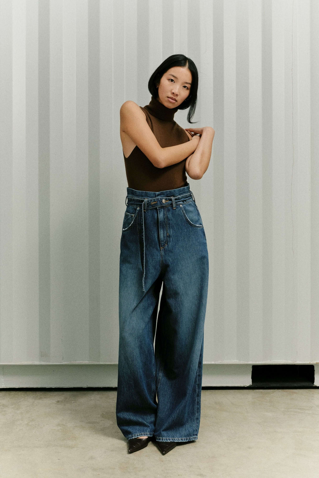 Eine Frau in einem ärmellosen braunen Rollkragen-Top und einer weit geschnittenen Jeans vor einem gestreiften Hintergrund.