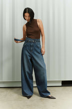 Eine Frau in einem braunen, ärmellosen Top hält den Bindegürtel ihrer weit geschnittenen Jeans in der Hand.