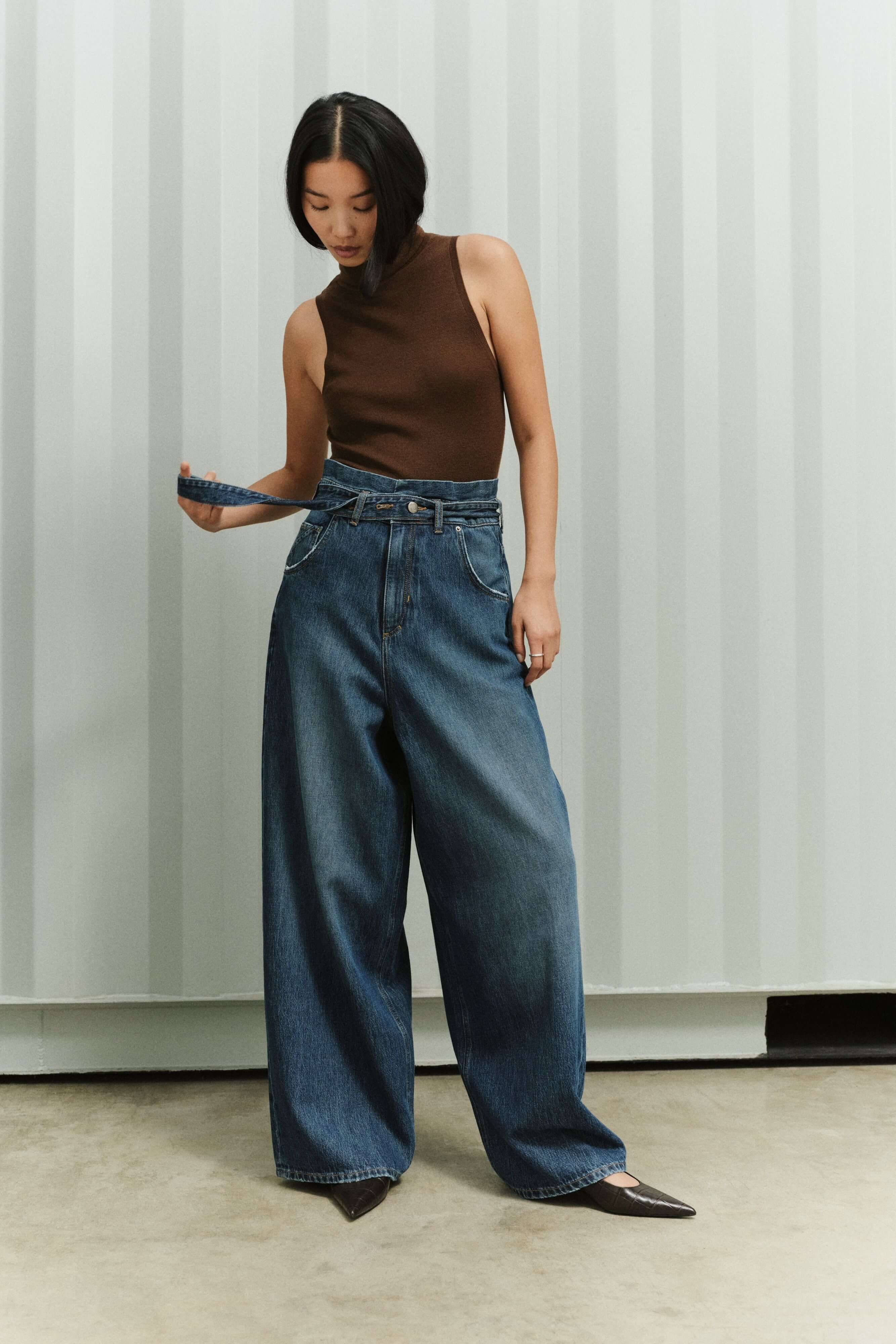 Eine Frau in einem braunen, ärmellosen Top hält den Bindegürtel ihrer weit geschnittenen Jeans in der Hand.