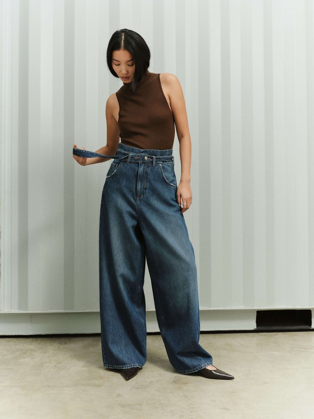 Frau in braunem, ärmellosem Top und blauer Jeans mit hohem Bund vor einer hellen, gestreiften Wand.
