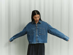 Frau in einer kastigen blauen Jeansjacke mit großen Taschen und dunkler Bundfaltenhose vor gestreiftem Hintergrund.