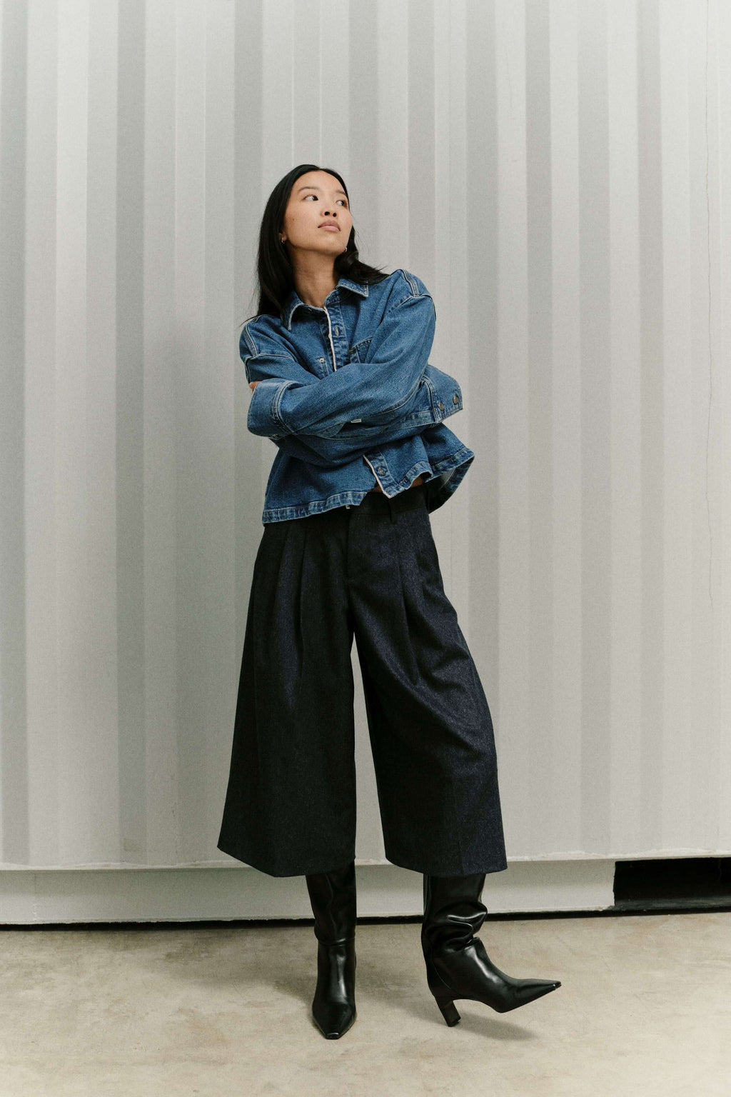 Frau in blauer Jeansjacke, dunkler Culotte mit weitem Bein und schwarzen Stiefeln vor gestreiftem Hintergrund.