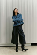 Frau in blauer Jeansjacke, dunkler Culotte mit weitem Bein und schwarzen Stiefeln vor gestreiftem Hintergrund.