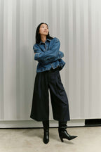 Frau in blauer Jeansjacke, dunkler Culotte mit weitem Bein und schwarzen Stiefeln vor gestreiftem Hintergrund.